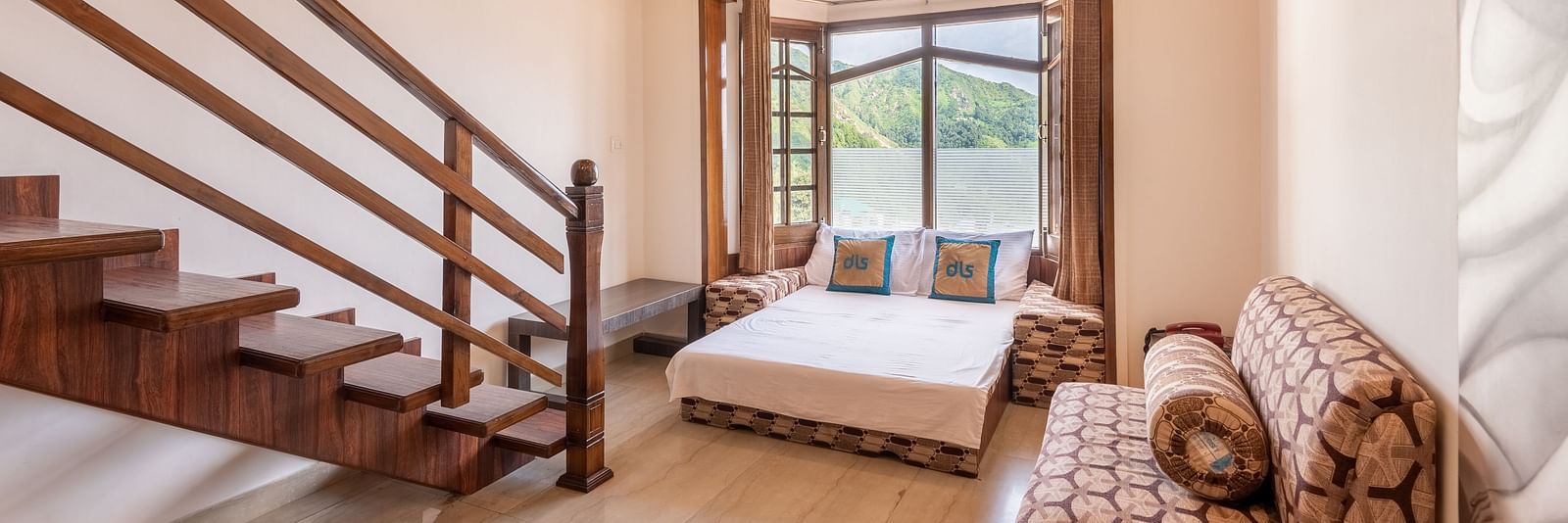 A spacious room near Bhagsunag Temple featuring a wooden staircase, a bed, a sofa, and a large window offering a mountain view – DLS Meghavan Resort.